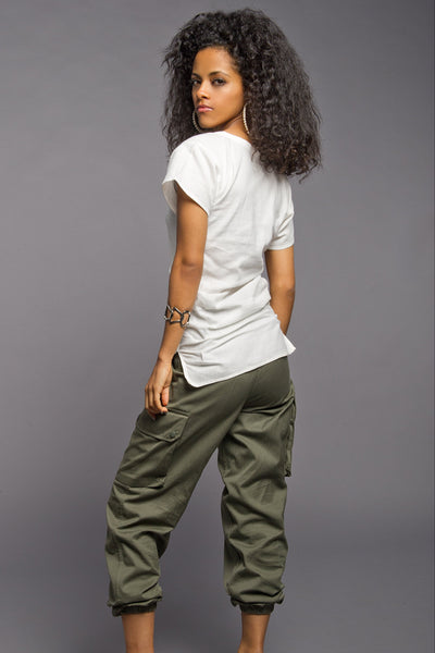 A woman with curly black hair is shown from a back-three-quarter angle, wearing a white short-sleeve t-shirt, olive green cargo pants, and brown high heels against a solid gray studio background.