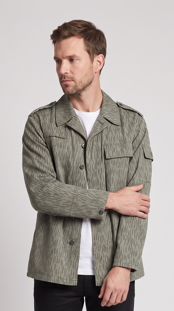 A man modeling a textured olive green utility jacket over a white t-shirt, black pants, and white sneakers, standing against a light gray studio background.
