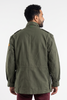 A man viewed from the back wearing an olive green military-style jacket and burgundy pants, standing against a plain white studio background.