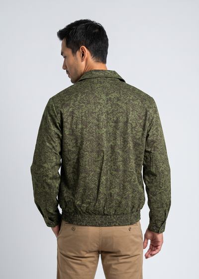 A man viewed from the back wearing an olive green patterned knit jacket and tan pants, standing against a light gray studio background.