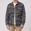 A man modeling a camouflage utility jacket with green, blue, and brown tones, paired with khaki pants and sneakers, against a light gray studio background.