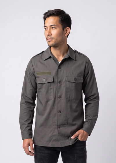 A man wearing a dark gray long-sleeve military-style shirt with flap chest pockets and dark jeans, standing against a light gray studio background.