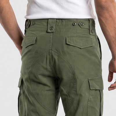 A man shown from the back wearing olive green cargo pants, a white tank top, and black sneakers against a light gray studio background.