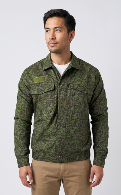 A man wearing a green camouflage long-sleeve button-up shirt over a white undershirt and khaki pants, standing against a light gray studio background.