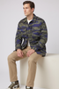 A man sits on a white block wearing a green and blue camouflage button-up shirt, khaki pants, and brown and white sneakers against a light gray studio background