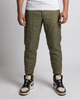 A man modeling olive green tapered cargo pants with a white tank top and black and white sneakers, standing against a light gray studio background.