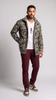 A man modeling a camouflage patterned jacket over a white t-shirt, paired with burgundy pants and brown sneakers, standing against a light gray studio background.