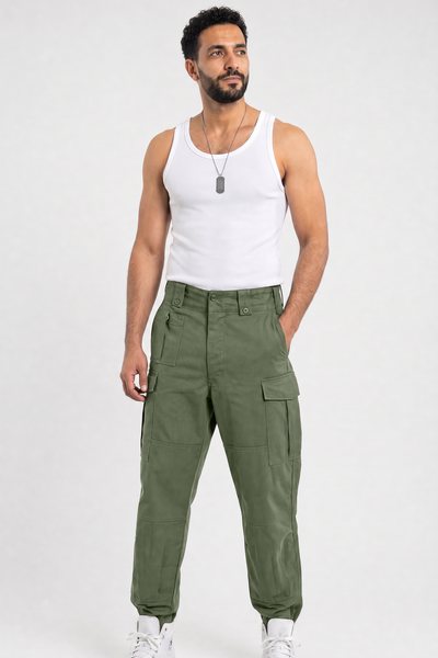 A man modeling olive green cargo pants with a white tank top and white sneakers, standing against a light gray studio background.