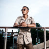 Man wearing sunglasses and a beige jacket standing on a bridge with buildings in the background