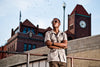 Person standing with arms crossed in front of a brick building with a clock tower.