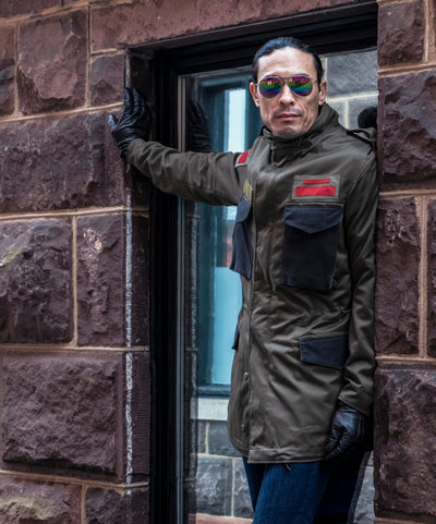 A man wearing a vintage olive green military-style jacket, sunglasses, and black gloves leans against a dark doorway framed by a brown stone wall. The 'TOP RANK VINTAGE' logo is visible in the top right corner.