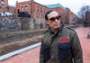 A man wearing a military-style olive green jacket with dark blue pockets and sunglasses, standing outdoors on a brick walkway with brick buildings in the background.