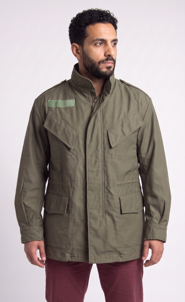 A man wearing an olive green military-style field jacket with a high collar and front pockets, paired with burgundy pants, photographed against a light gray studio background.