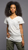 A woman with voluminous dark curly hair wearing a white short-sleeve V-neck t-shirt and olive green pants, standing against a solid gray studio background.