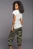 A woman with curly black hair is shown from a back-three-quarter angle, wearing a white short-sleeve t-shirt, olive green cargo pants, and brown high heels against a solid gray studio background.