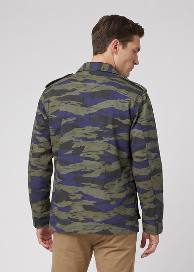 A man viewed from the back wearing a camouflage jacket with olive green, blue, and dark tones, paired with tan trousers and brown shoes, against a light gray studio background.