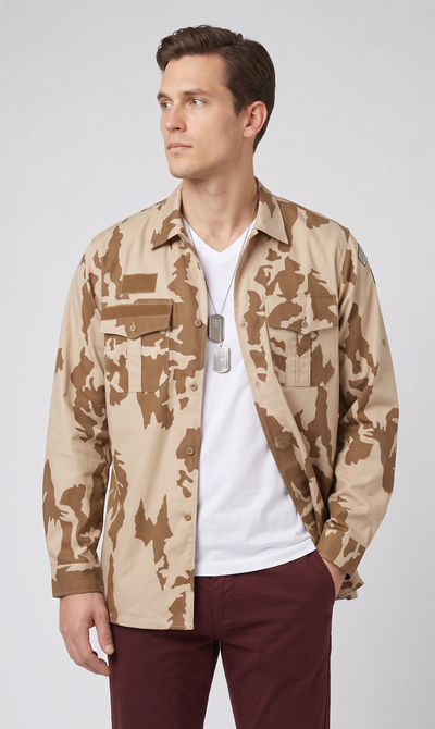 A man wearing a desert camouflage button-up shirt over a white t-shirt and dark burgundy pants, standing against a light gray studio background.