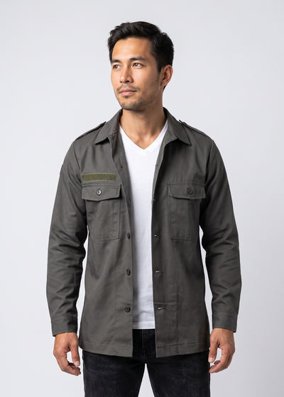 A man wearing a dark olive green utility field jacket over a white t-shirt and black jeans, standing against a light gray studio background.
