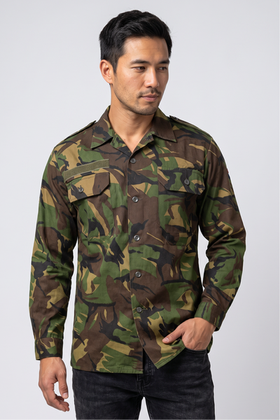 A man wearing a long-sleeve camouflage button-up shirt and dark jeans, standing against a light gray studio background.