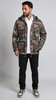 A man wearing a camouflage hooded jacket over a white shirt, black pants, and sneakers, standing against a light gray studio background.