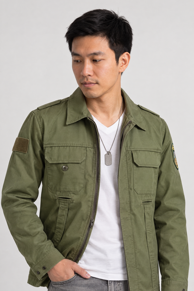 A man wearing an olive green military-style utility jacket over a white t-shirt and gray jeans, standing against a light gray studio background
