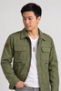 A man wearing an olive green military-style utility jacket over a white t-shirt and gray jeans, standing against a light gray studio background