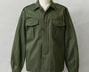 An olive green military-style field jacket with a pointed collar, button front, and two large chest flap pockets, displayed on a mannequin torso against a light gray textured background.