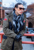 A man wearing an olive green military-style field coat, a gray and black plaid scarf, black gloves, and sunglasses, leaning on a red railing in an outdoor urban setting.