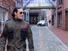 A man wearing a military-style olive green utility jacket with dark blue pockets walks down a cobblestone alleyway lined with brick buildings.