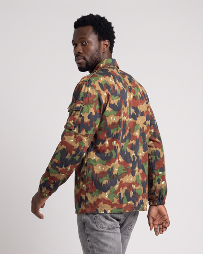 A man wearing a camouflage utility jacket and gray jeans, shown from a side-back angle against a light gray studio background.
