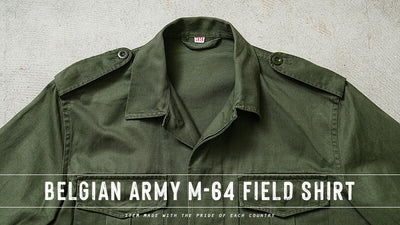 An olive green military-style field shirt laid flat against a light textured background, with a white text overlay at the bottom reading 'BELGIAN ARMY M-64 FIELD SHIRT'.