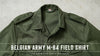 An olive green military-style field shirt laid flat against a light textured background, with a white text overlay at the bottom reading 'BELGIAN ARMY M-64 FIELD SHIRT'.